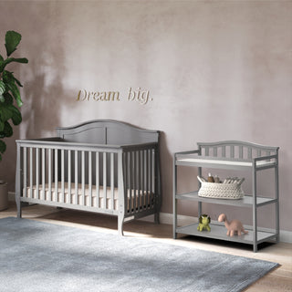 A Child Craft Camden 4-in-1 Convertible Crib in Cool Grey with a matching changing table, toys, and basket sits by a light pink wall with "Dream big," while a potted plant and blue rug add to the cozy nursery.