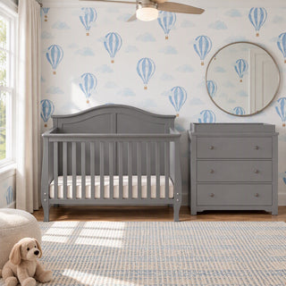 A grey wooden Camden 2-Piece Nursery Set With Crib and Dresser stands against a wall with blue hot air balloon wallpaper, featuring a 3-drawer dresser with a changing table topper.