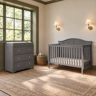 A gray Child Craft Camden 2-Piece Nursery Set With Crib and Dresser consisting of a convertible crib and a matching three-drawer dresser with a changing table topper.