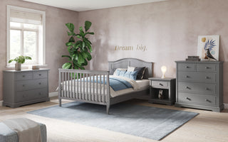 A bright bedroom with light pink walls, gray wooden furniture, a Child Craft Camden 4-in-1 Convertible Crib in Cool Grey, a bed with blue and white bedding, dresser, nightstand, chest of drawers, a large green plant, rug, and “Dream big” on the wall.