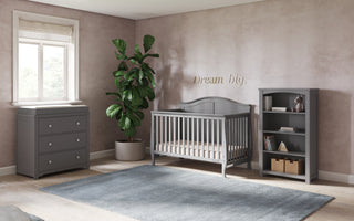 A modern nursery with mauve walls, a window with a Roman shade, a potted plant, a blue rug, "Dream big" above the crib, and a Child Craft Camden 4-in-1 Convertible Crib in Cool Grey with matching dresser and bookshelf.