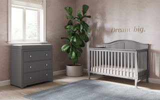 A softly lit nursery features the Child Craft Harmony 3-Drawer Dresser with Changing Table Topper in cool gray, a gray crib, large potted plant, gray rug, and "Dream big." written in gold on the pinkish wall near a window.