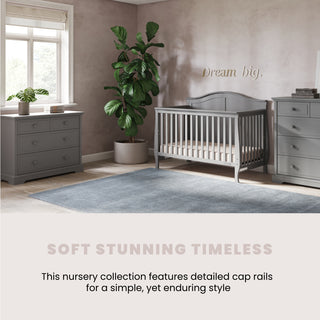 A peaceful nursery features Child Craft’s Camden 4-in-1 Convertible Crib in Cool Grey, matching furniture, a plant by the window, and “Dream big.” above the crib. The room has a soft glow and tranquil vibe.