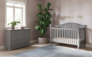 A cozy nursery with a large window, gray dresser, potted plant, and the Child Craft Camden 4-in-1 Convertible Crib in Cool Grey sits on a blue rug. “Dream big.” in gold decorates the wall as soft natural light fills the room.