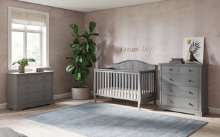 A cozy nursery features gray furniture, including the Child Craft Camden 4-in-1 Convertible Crib in Cool Grey, a dresser, and chest of drawers. A potted plant sits by the window, with "Dream big" written on the pink wall above the crib.