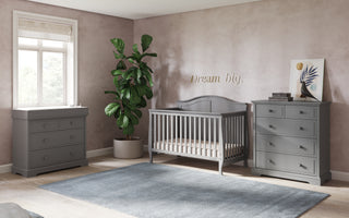 A nursery with gray furniture, featuring the Child Craft Camden 4-in-1 Convertible Crib in Cool Grey, a matching dresser and changing table, blue rug, potted plant, “Dream big” wall art, and a window with a Roman shade letting in light.