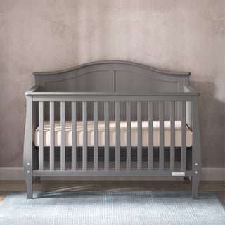 The Child Craft Camden 4-in-1 Convertible Crib in Cool Grey, featuring vertical slats and a curved headboard, stands on a blue rug against a light pink textured wall. It includes a beige mattress for long-lasting comfort and style.