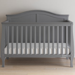 A Child Craft Camden 4-in-1 Convertible Crib in Cool Grey with slatted sides and a curved headboard sits on a light wood floor against a plain light wall, featuring a simple white mattress.
