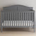 A Child Craft Camden 4-in-1 Convertible Crib in Cool Grey with slatted sides and a curved headboard sits on a light wood floor against a plain light wall, featuring a simple white mattress.