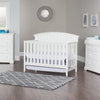 A nursery with white furniture, including a crib, dresser with the Child Craft F01792 Universal Changing Table Topper in Matte White, sits on a gray wood floor and patterned rug. Gray walls feature a round mirror and framed art.