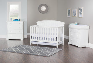 A nursery with white furniture, including a crib, dresser with the Child Craft F01792 Universal Changing Table Topper in Matte White, sits on a gray wood floor and patterned rug. Gray walls feature a round mirror and framed art.