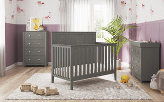 A nursery features the Child Craft Atwood 4-in-1 Convertible Crib in Lunar Gray, matching dresser and changing table on a light rug. Toys scatter the floor, a potted plant sits nearby, and sunlight streams through curtained windows onto playful purple wallpaper.