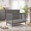 A cozy nursery with a gray crib, Child Craft dresser featuring the Universal Changing Table Topper in Lunar Gray, a plush rug, wooden toys, and a plant by the window. Light pink wallpaper and purple wainscoting adorn the walls.