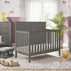 A cozy nursery features the Child Craft Atwood 4-in-1 Convertible Crib in Lunar Gray, matching gray furniture, and a soft rug. Toys and stuffed animals lie on the floor, while a plant and lamp add warmth by a window with sheer curtains.