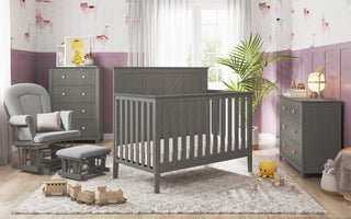A cozy nursery features the Child Craft Atwood 4-in-1 Convertible Crib in Lunar Gray, matching gray furniture, and a soft rug. Toys and stuffed animals lie on the floor, while a plant and lamp add warmth by a window with sheer curtains.