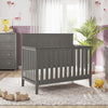 A modern nursery featuring the Child Craft Atwood 4-in-1 Convertible Crib in Lunar Gray, matching dressers, plush rug, toy train, stuffed animals, and potted plant. Sunlight streams through sheer curtains over animal-themed wallpaper.