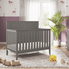 The Child Craft Atwood 4-in-1 Convertible Crib in Lunar Gray sits on a patterned rug in a softly lit nursery with mauve paneled walls, a dresser, plush toys, wooden train, blocks, potted plant, and sheer-curtained window.