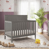 The Child Craft Atwood 4-in-1 Convertible Crib in Lunar Gray sits on a patterned rug in a nursery with purple wainscoting, pink wallpaper, wooden toys, a yellow plush duck, dresser, plant, and a window with sheer curtains in the background.