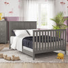 A cozy bedroom features the Child Craft Atwood 4-in-1 Convertible Crib in Lunar Gray, gray furniture, a dresser, chest, a large window with sheer curtains, wooden toys on a gray rug, and light purple walls with playful wallpaper accents.