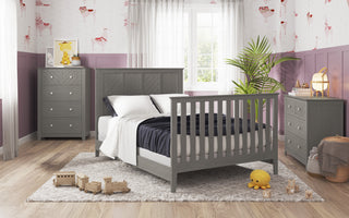 A cozy bedroom features the Child Craft Atwood 4-in-1 Convertible Crib in Lunar Gray, gray furniture, a dresser, chest, a large window with sheer curtains, wooden toys on a gray rug, and light purple walls with playful wallpaper accents.