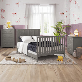 A cozy, modern kids bedroom featuring the Child Craft Atwood 4-in-1 Convertible Crib in Lunar Gray, with a matching dresser and nightstand, plush toys, a plant, and natural light from a large window with sheer curtains.