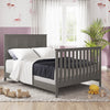 A cozy bedroom featuring the Child Craft Atwood 4-in-1 Convertible Crib in Lunar Gray, soft white bedding, purple walls, wooden toys, a plush octopus, a dresser, a plant, and sheer curtains filtering natural light.