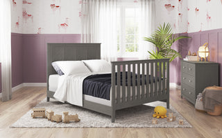 A cozy bedroom featuring the Child Craft Atwood 4-in-1 Convertible Crib in Lunar Gray, soft white bedding, purple walls, wooden toys, a plush octopus, a dresser, a plant, and sheer curtains filtering natural light.