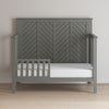 The Child Craft Atwood 4-in-1 Convertible Crib in Lunar Gray features a chevron-patterned wooden headboard and partial guard rail. Shown against a light wall on a light wood floor with a bare white mattress.