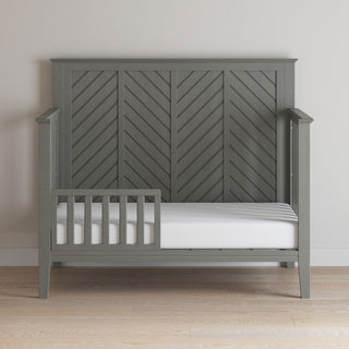 The Child Craft Atwood 4-in-1 Convertible Crib in Lunar Gray features a chevron-patterned wooden headboard and partial guard rail. Shown against a light wall on a light wood floor with a bare white mattress.