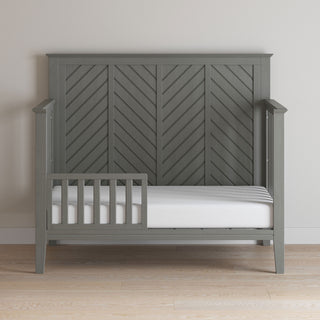 The Child Craft F09501 Toddler Guard Rail in Lunar Gray features sturdy wood construction, a chevron headboard, three open sides, and a partial safety rail on one side, set on a light wood floor against a plain light wall.