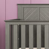 A close-up of the Child Craft Atwood 4-in-1 Convertible Crib in Lunar Gray, featuring vertical slats and a modern chevron back panel, set against a pink paneled wall.