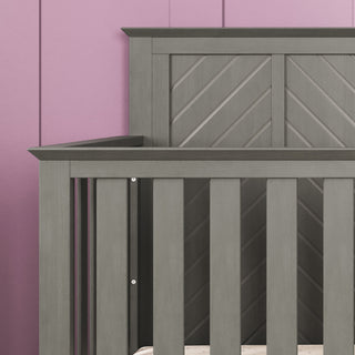 A close-up of the Child Craft Atwood 4-in-1 Convertible Crib in Lunar Gray, featuring vertical slats and a modern chevron back panel, set against a pink paneled wall.