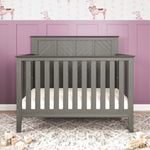 The Child Craft Atwood 4-in-1 Convertible Crib in Lunar Gray stands against a mauve paneled wall with playful llama wallpaper, while wooden alphabet blocks are scattered on the light floor in front of the crib.
