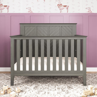 The Child Craft Atwood 4-in-1 Convertible Crib in Lunar Gray stands against a mauve paneled wall with playful llama wallpaper, while wooden alphabet blocks are scattered on the light floor in front of the crib.