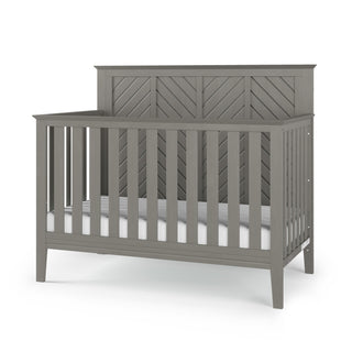 The Child Craft Atwood 4-in-1 Convertible Crib in Lunar Gray features a high chevron-patterned back panel, vertical side and front slats, four sturdy legs, and comes with a white mattress.