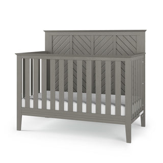 The Child Craft Atwood 4-in-1 Convertible Crib in Lunar Gray features a high chevron-patterned back panel, vertical side and front slats, four sturdy legs, and comes with a white mattress.