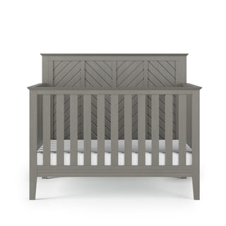 The Child Craft Atwood 4-in-1 Convertible Crib in Lunar Gray features vertical slats, a chevron-patterned headboard, and a white mattress, all set against a plain white background.