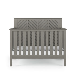 The Child Craft Atwood 4-in-1 Convertible Crib in Lunar Gray features vertical slats, a chevron-patterned headboard, and a white mattress, all set against a plain white background.