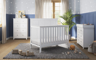 A bright nursery features the Child Craft Atwood 4-in-1 Convertible Crib in matte white, matched with a dresser and changing table on a gray rug. Wooden toys, a teddy bear, a large plant, and star wallpaper add charm to the blue and white walls.