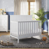A bright nursery with a white crib, dresser, and the Child Craft F01792 Universal Changing Table Topper in Matte White. Toys and stuffed animals sit on a gray rug. Sunlight shines on blue star-patterned walls and a large plant by the crib.