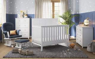 Bright nursery with a white crib, dresser, chest, gray rug, plush toys, wooden train, potted plant, star-patterned wallpaper, and natural light. Features the Child Craft Tranquil Nursery Glider and Ottoman in Matte White for cozy seating.
