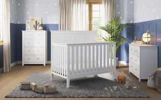 A nursery features the Child Craft Atwood 4-in-1 Convertible Crib in Matte White, a matching changing table and dresser on a gray rug. Wooden toys and a plush turtle rest on the floor, with blue and gray star wallpaper and natural light from the window.