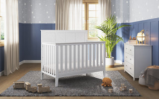 A bright nursery features the Child Craft Atwood 4-in-1 Convertible Crib in matte white, centered on a gray rug, with wooden toys, a lamp-topped dresser, a large potted plant, and star-patterned walls with blue paneling and sunny windows.