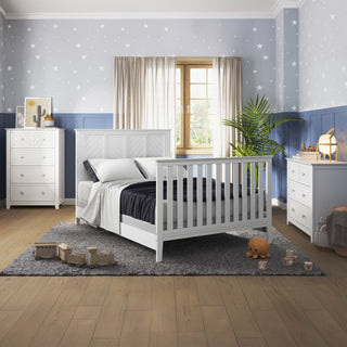A cozy kids’ bedroom featuring the Child Craft Atwood 4-in-1 Convertible Crib in matte white, a gray rug, star-patterned wallpaper, two white dressers, wooden toys on the floor, a potted plant, and soft natural light through beige curtains.