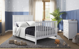 A cozy children’s room features the Child Craft Atwood 4-in-1 Convertible Crib in Matte White, navy and white bedding, a dresser with changing pad, toys on a gray rug, and blue star-patterned wallpaper. Sunlight streams through beige-curtained windows.