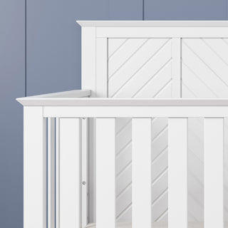 A close-up of the Child Craft Atwood 4-in-1 Convertible Crib in Matte White, featuring vertical slats and a chevron-patterned panel, set against a blue wall with vertical grooves.