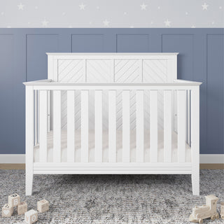 The Child Craft Atwood 4-in-1 Convertible Crib in Matte White, featuring a chevron headboard, stands on a gray textured rug with wooden alphabet blocks. Behind it is a wall of blue paneling and white star-patterned wallpaper.
