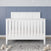 The Child Craft Atwood 4-in-1 Convertible Crib in Matte White, featuring a chevron headboard, stands on a gray textured rug with wooden alphabet blocks. Behind it is a wall of blue paneling and white star-patterned wallpaper.
