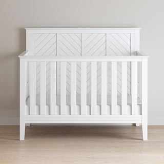 The Child Craft Atwood 4-in-1 Convertible Crib in Matte White features a chevron-patterned headboard and vertical side slats, shown here without bedding on a light wood floor against a plain beige wall.