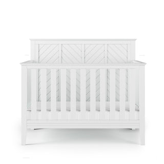 The Child Craft Atwood 4-in-1 Convertible Crib in Matte White features vertical slats and a decorative chevron-patterned headboard, displayed from the front against a white background.